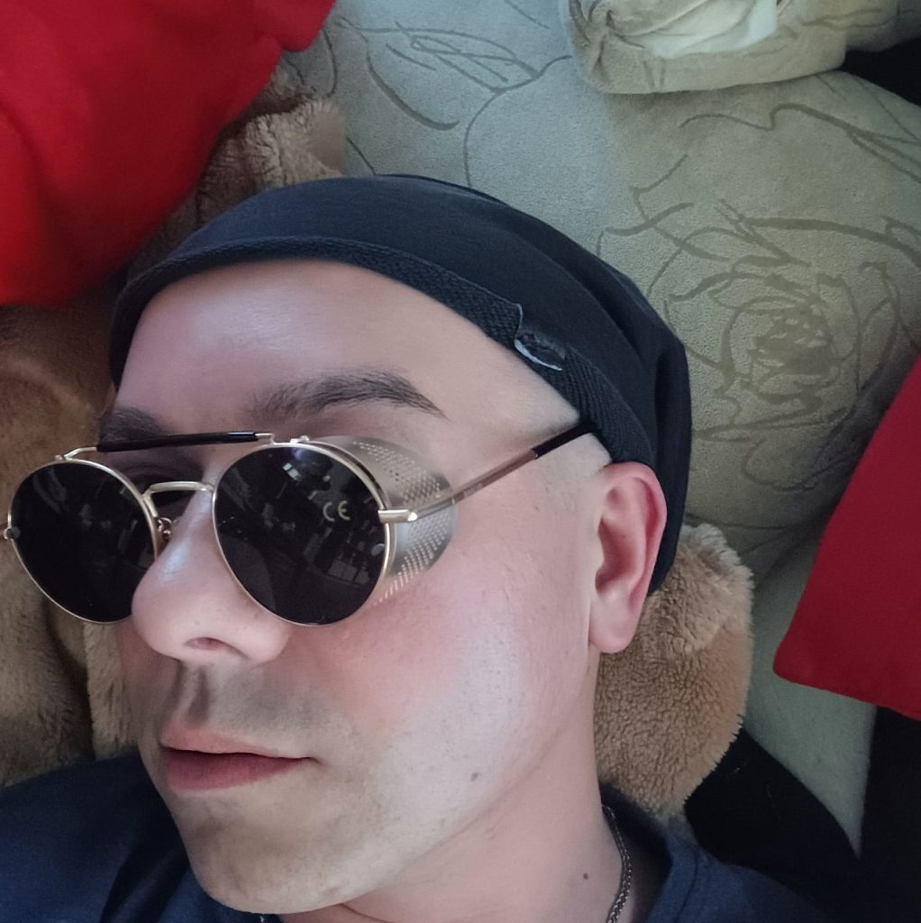 Zbliżenie na mężczyznę leżącego na pluszowych poduszkach. Ma na głowie czarną beanie i nosi okrągłe, ciemne okulary przeciwsłoneczne z bocznymi osłonami w stylu steampunkowym. Ma na sobie ciemną koszulkę, a w tle widać poduszki w beżowych i czerwonych odcieniach.